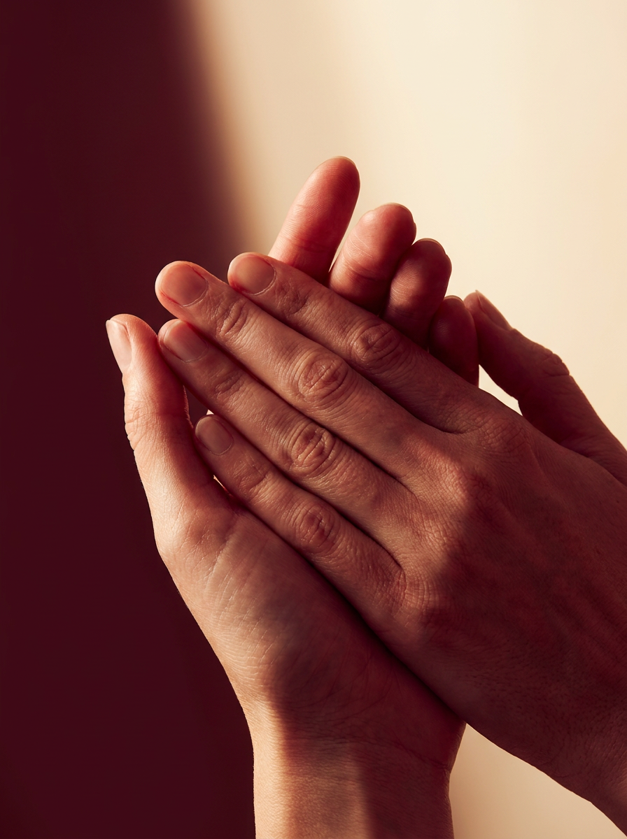 Two hands pressed together in warm light against a deep red backdrop.